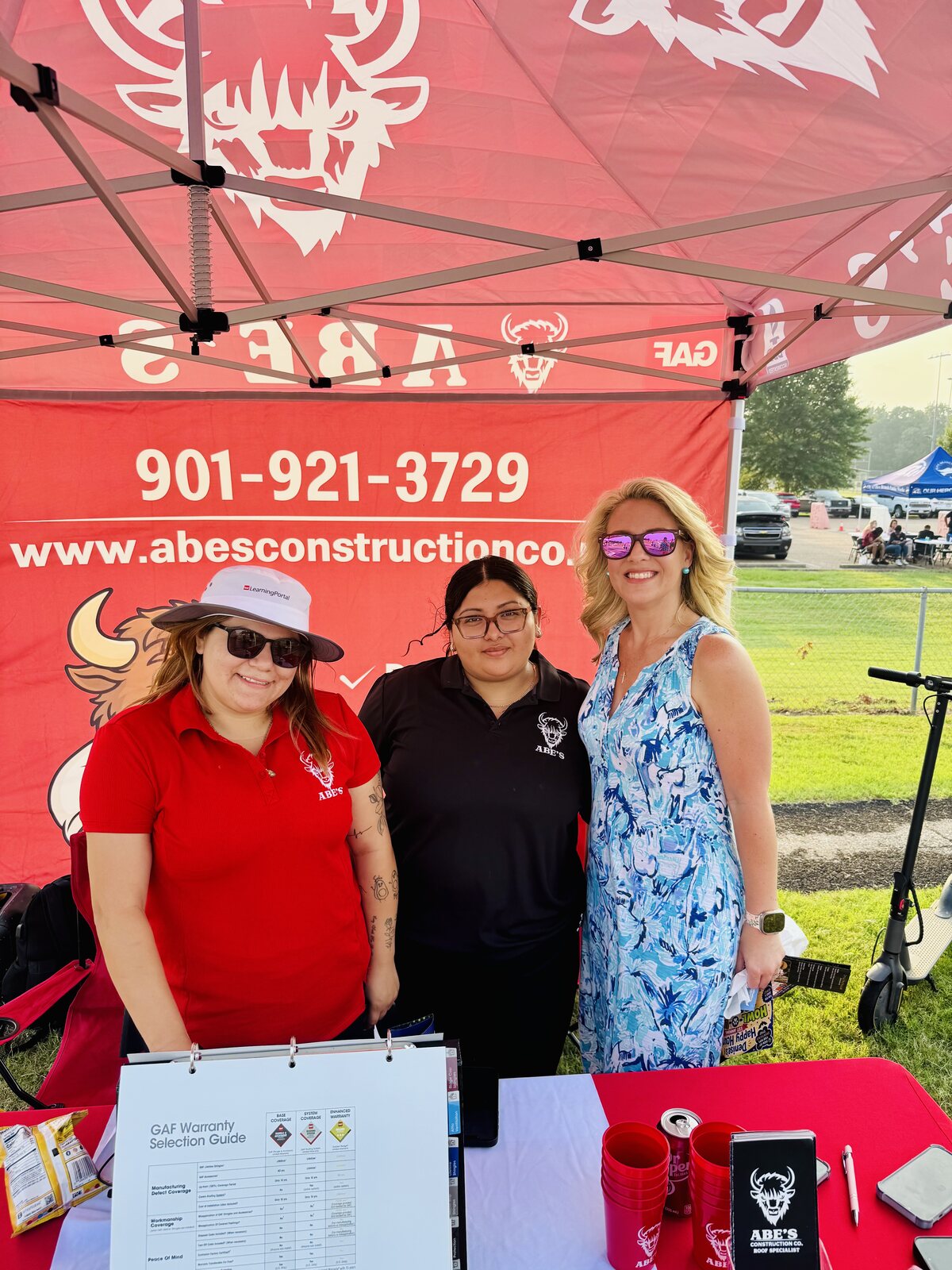 Three people posing at Abe's booth beneath a red canopy at the Olive Branch community day