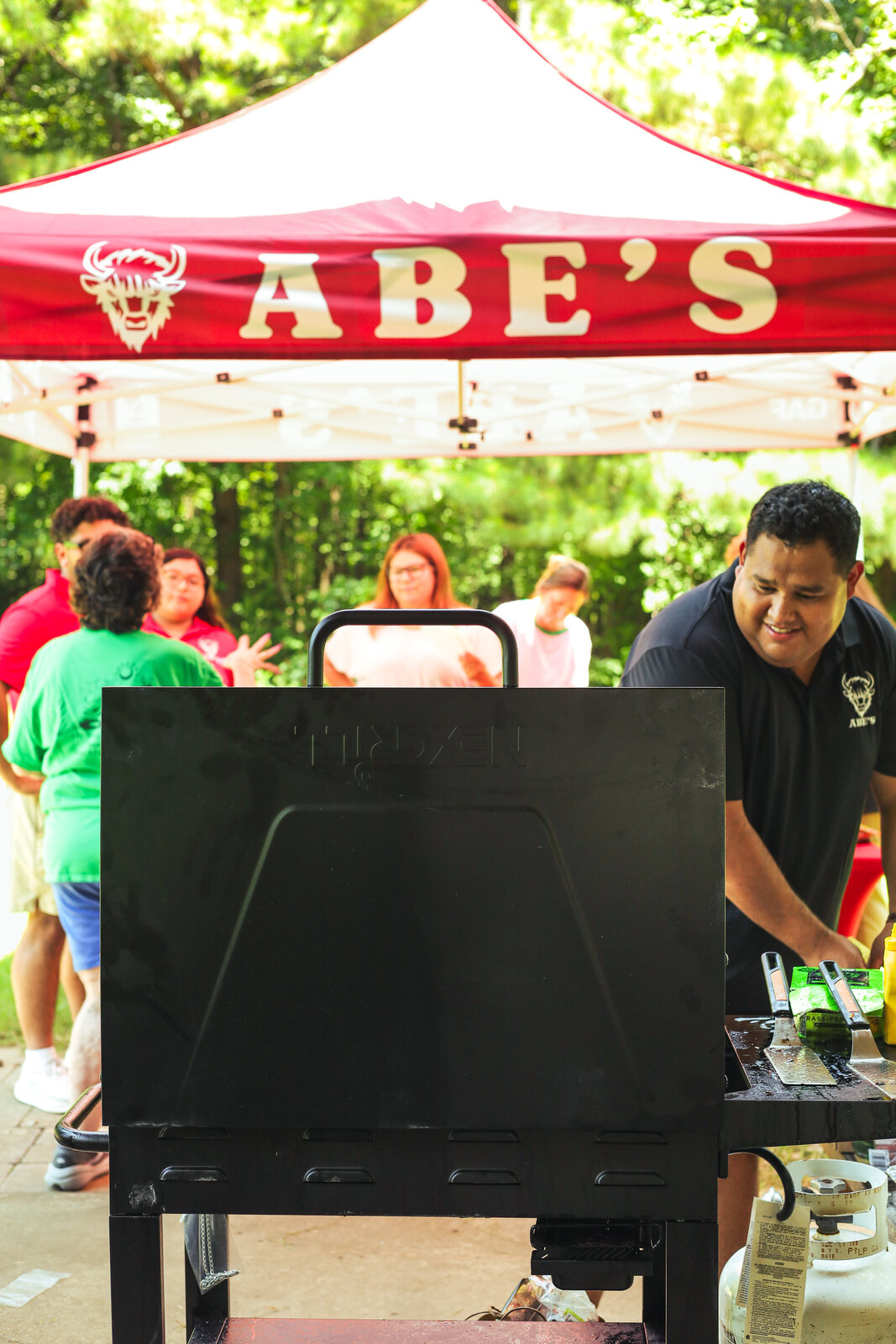 Abe's canopy behind the outdoor grill as people gather nearby